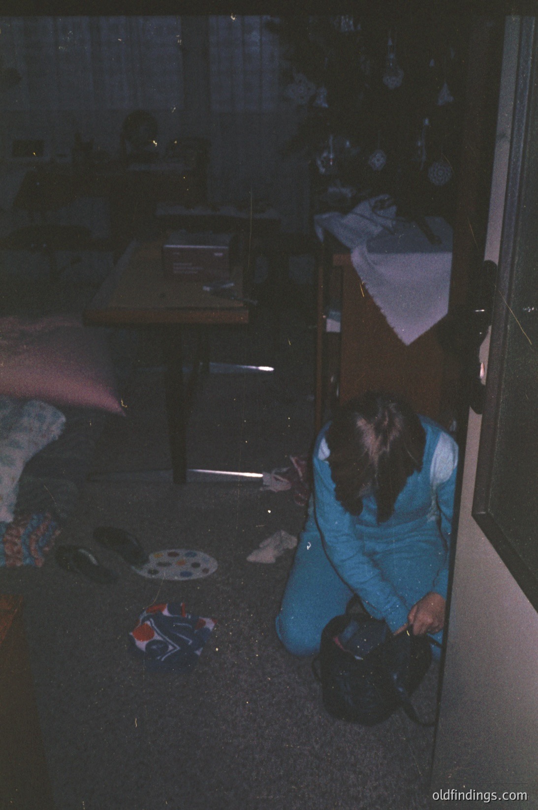 Vintage interior shot of a dimly lit bedroom with mid-century furnishings. A person in blue track pants kneels on patterned linoleum, holding a bucket near a sink. Wooden cabinetry and a bed with floral bedding suggest 1970s domestic life. Low-angle lighting enhances shadows.
