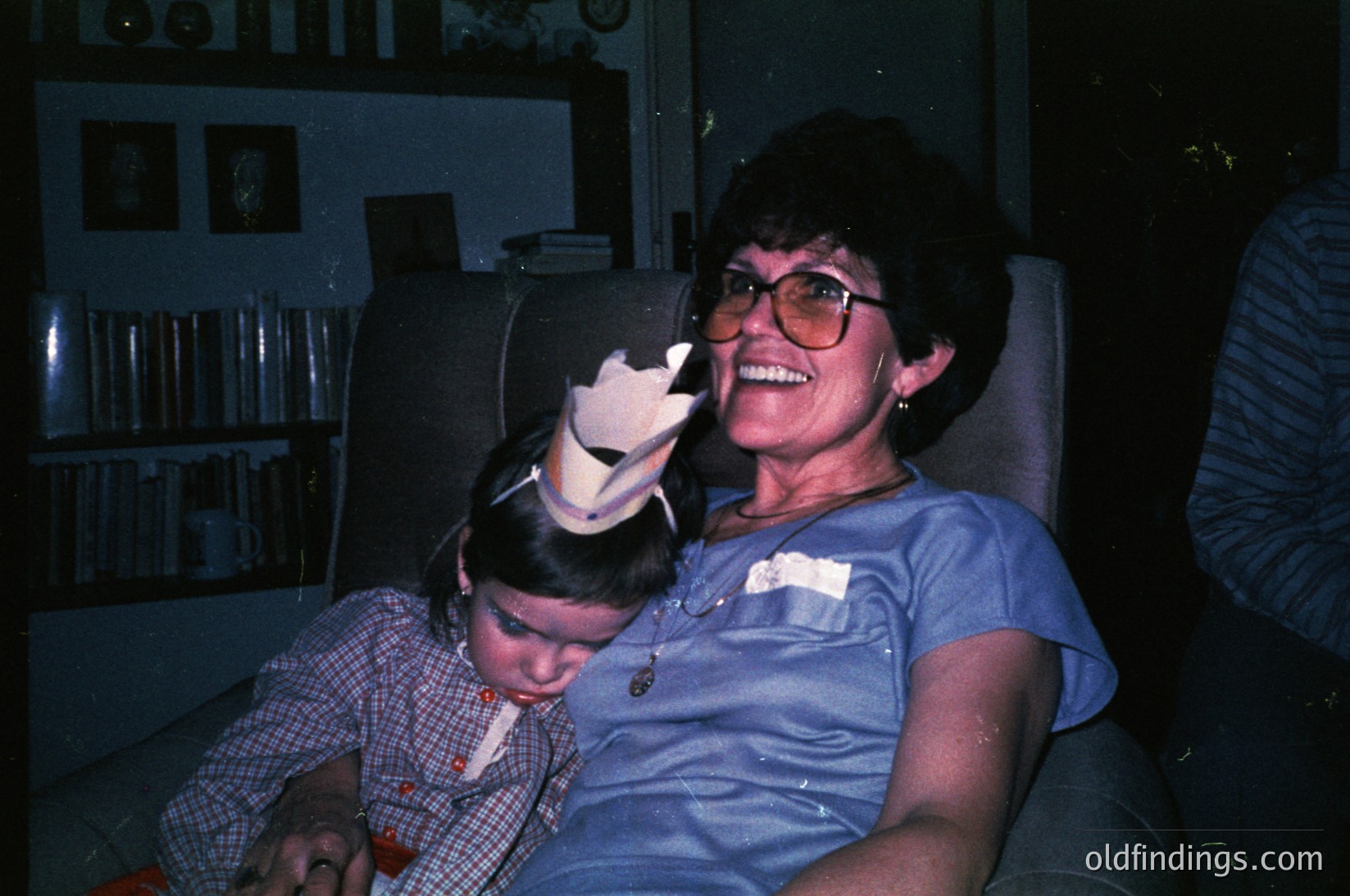 Vintage indoor portrait of a woman in a blue blouse and glasses holding a laughing child in a plaid shirt, likely from the 1970s–1980s. Warm lighting highlights framed photos and bookshelves in the background. Candid, nostalgic family moment.