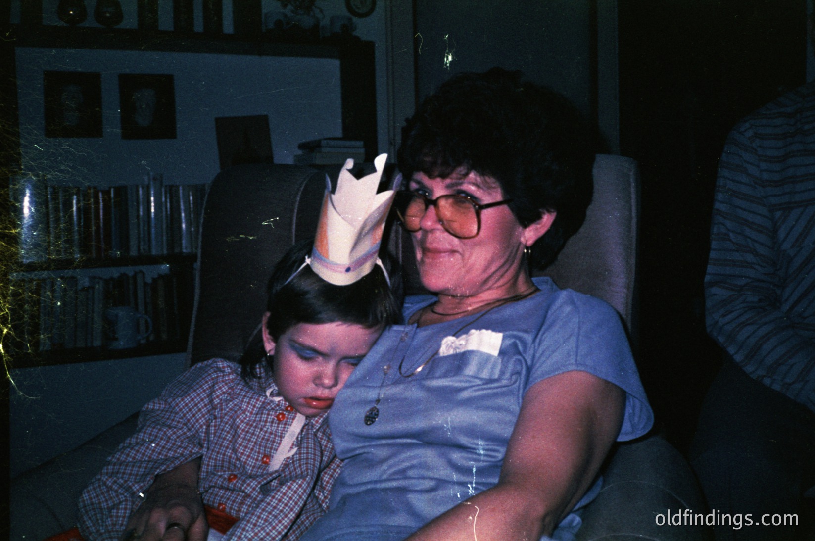 A grandmother and child share a tender moment indoors, likely during a birthday celebration. The woman wears a blue blouse, round glasses, and a crown, while the child dons a matching crown and plaid shirt. The dimly lit room features a bookshelf and a striped curtain, suggesting a mid-20th century home setting. [Intimate family moment with birthday crowns in a vintage home setting ]
