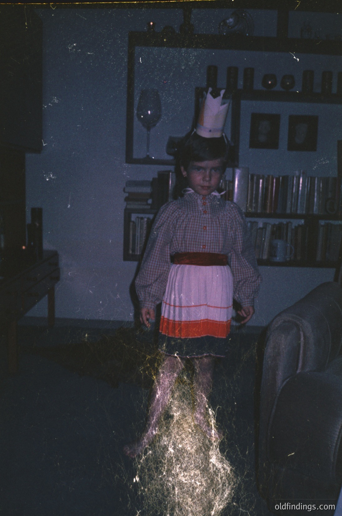 Vintage indoor portrait of a child in a princess dress with a crown, holding a sparkler. Warm lighting and blurred motion capture dynamic energy. Bookshelves and a wine glass in background suggest a festive, mid-20th-century home setting.