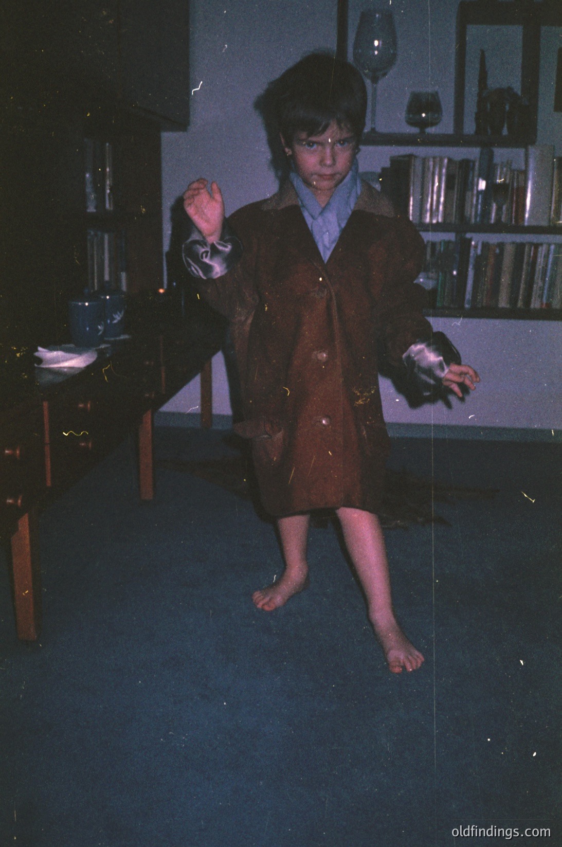 Young boy in vintage 1970s-80s suit, barefoot, mid-dance pose indoors. Bookshelf with glassware and books in background. Warm sepia tone suggests aged photo.