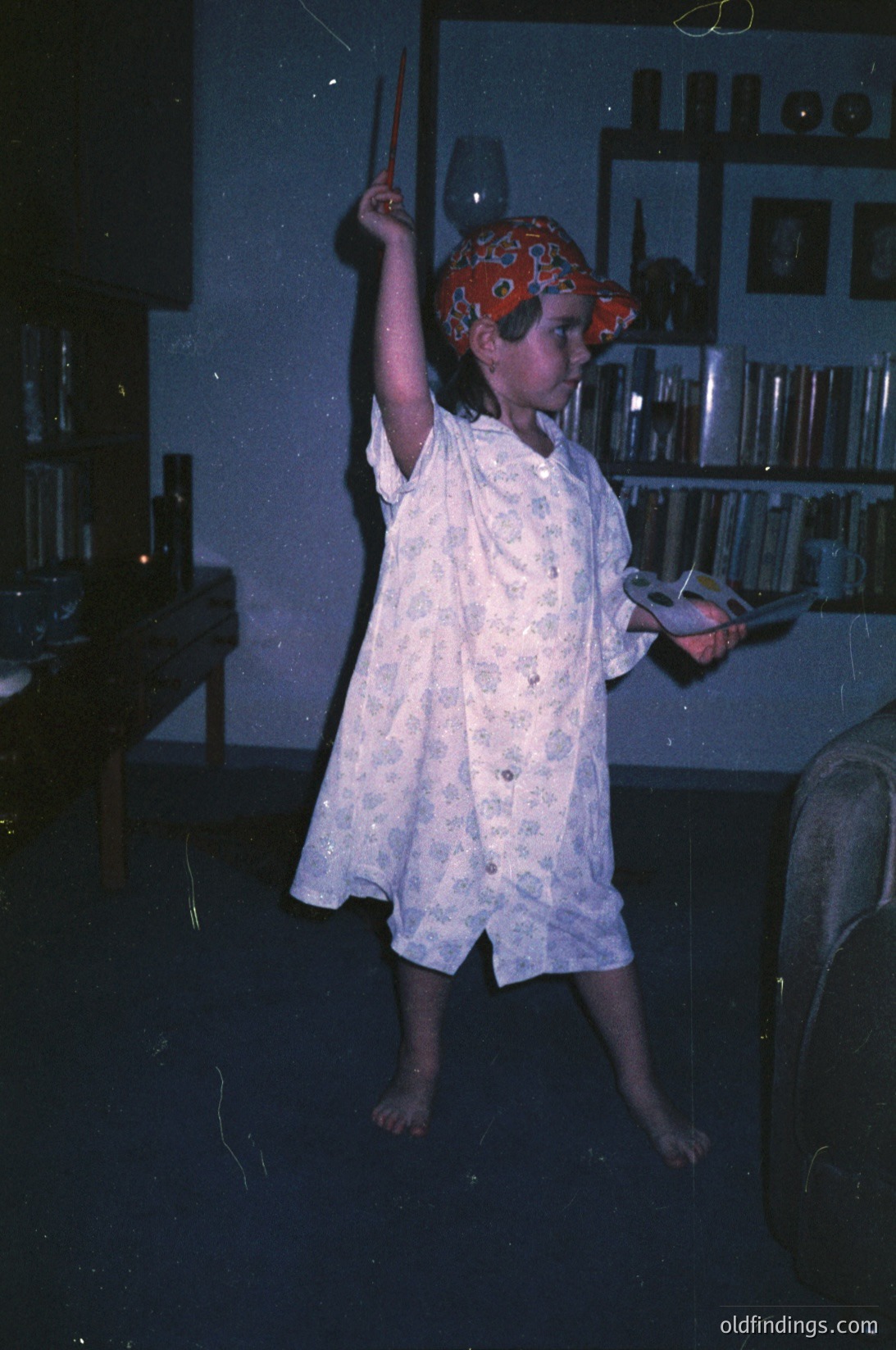 Child in 1970s-style pirate costume (red bandana hat, white floral robe) mid-motion indoors, holding a toy sword. Dimly lit room with bookshelves, vintage furniture, and a framed photo on the wall. Evokes mid-century home nostalgia.