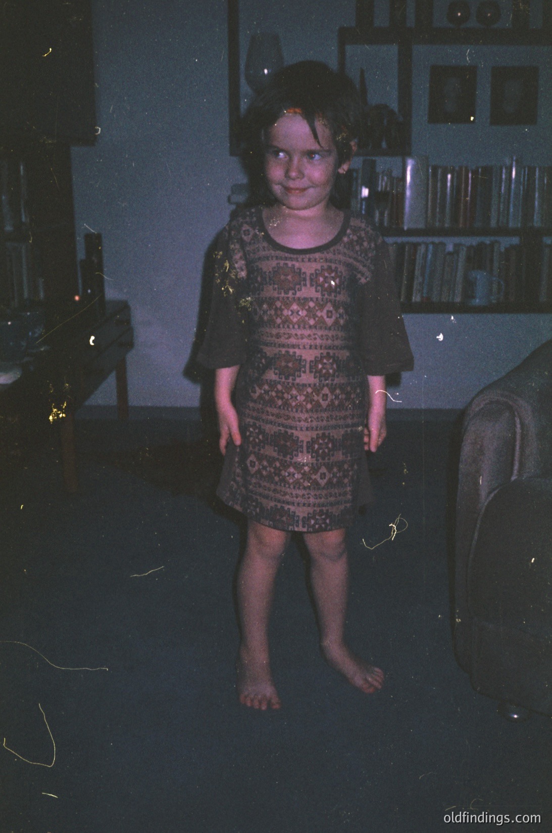 Vintage indoor portrait of a young girl in a patterned, knee-length dress, standing barefoot in dim lighting. Bookshelves with framed photos and a fireplace in the background suggest a cozy, lived-in home. Likely 1970s–1980s American suburban setting.