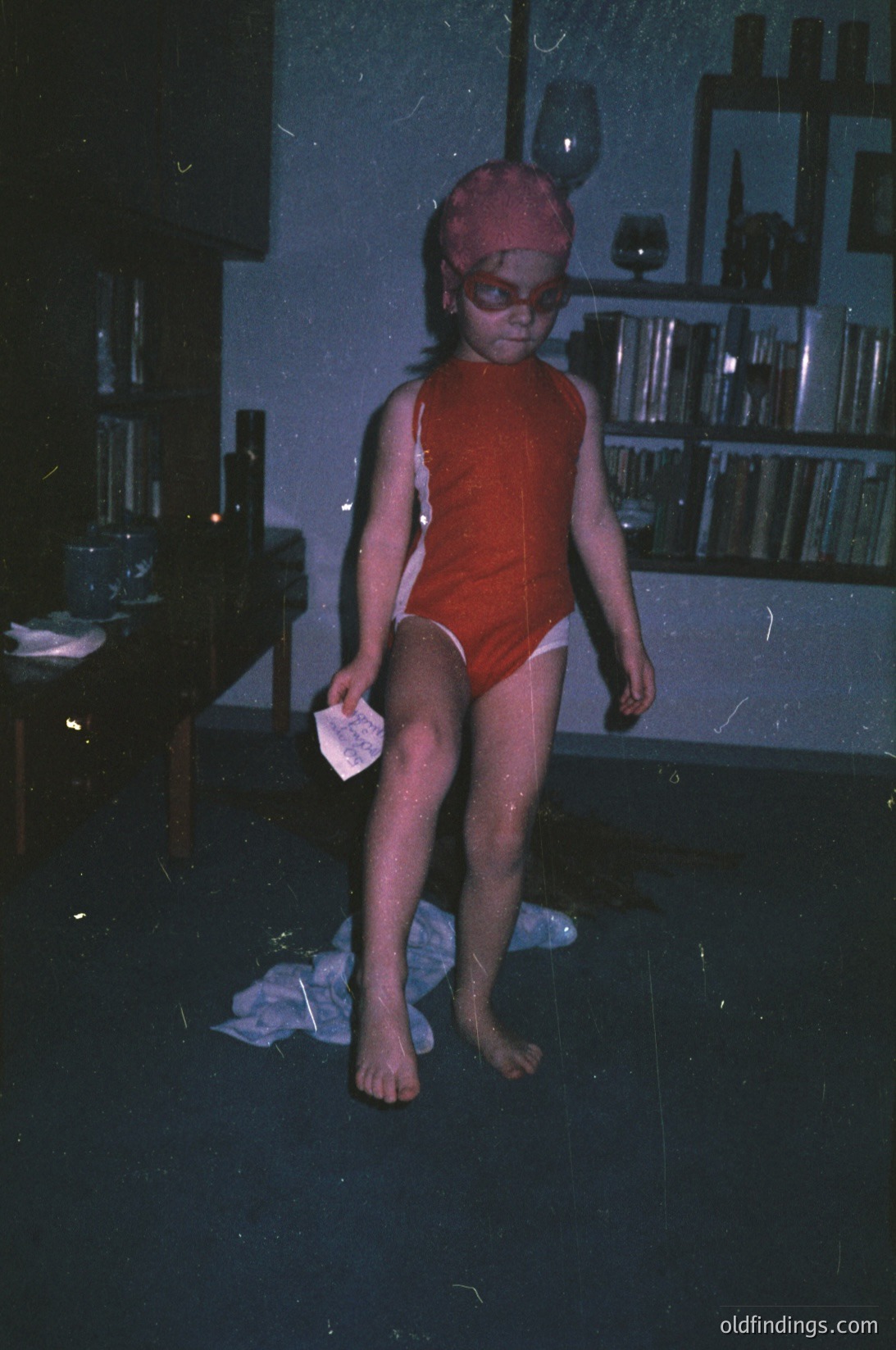 Child in vintage red swimwear and goggles stands indoors, likely a poolside changing area. Notebooks and glassware on shelves in background suggest a residential or small pool club setting. Color saturation and grain indicate a 1960s–1970s photo.