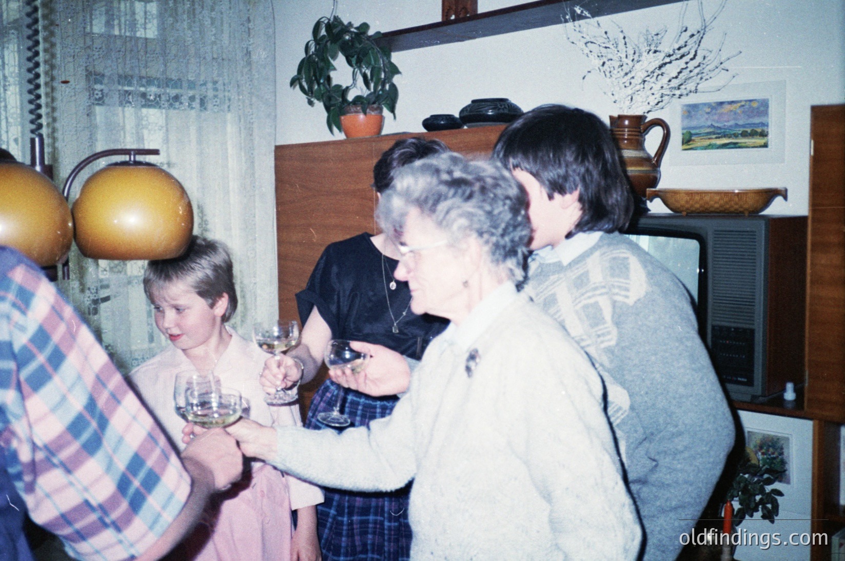Family gathering in a mid-century interior, likely 1970s–1980s. Three adults and a child stand in a living room with floral wallpaper, wooden furniture, and potted plants. The woman in the center holds a wineglass; the man beside her wears a patterned sweater. Decor includes a ceramic jug, framed artwork, and a hanging pumpkin. Warm, intimate setting.