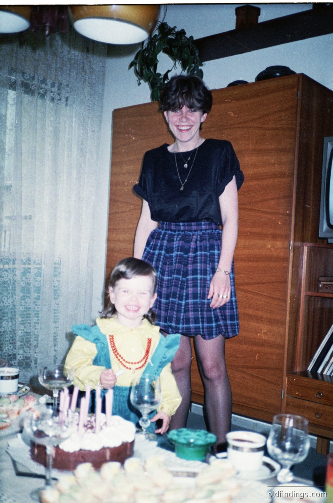 Vintage indoor birthday scene: woman in 1970s-style navy blouse & plaid skirt poses with young girl in yellow top & red beaded necklace beside a tiered chocolate cake. Wooden cabinet, floral curtains, and glassware suggest a mid-century home setting.