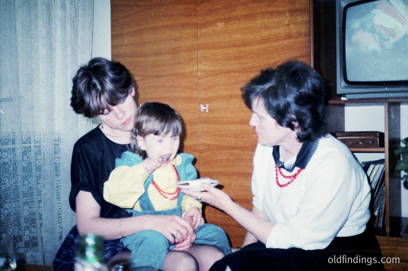 Vintage indoor family moment: two adults and a child seated on a sofa, sharing a snack. The woman wears a white blouse with red bead necklace, the teen a black top; child in a yellow shirt with green pants. A CRT TV plays in the background. Wood-paneled walls and a bookshelf suggest mid-20th century home decor.
