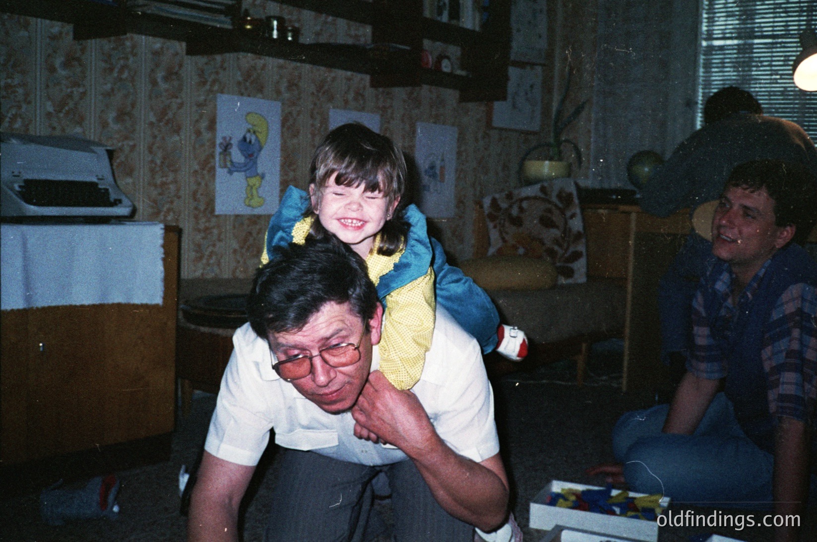 Vintage indoor family moment: man in glasses carries a smiling child in a yellow sweater, another adult watches. Patterned wallpaper, framed illustrations, and a globe on shelves suggest mid-20th-century home decor. Warm, candid lighting captures playful interaction.