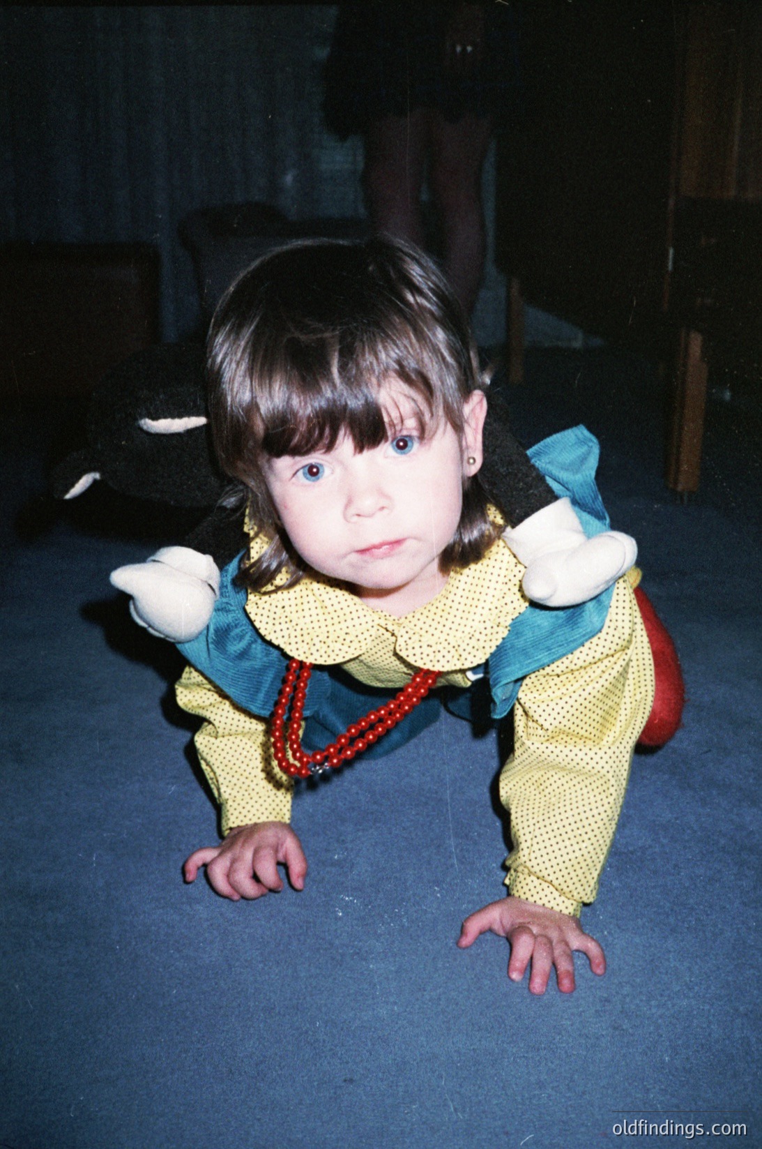 Young child in vintage-style outfit (yellow knit sweater with green collar, red beaded necklace) crawling on dark floor, holding a white stuffed animal. Indoor setting with wooden furniture and soft lighting, suggesting late 20th-century home decor ( ).