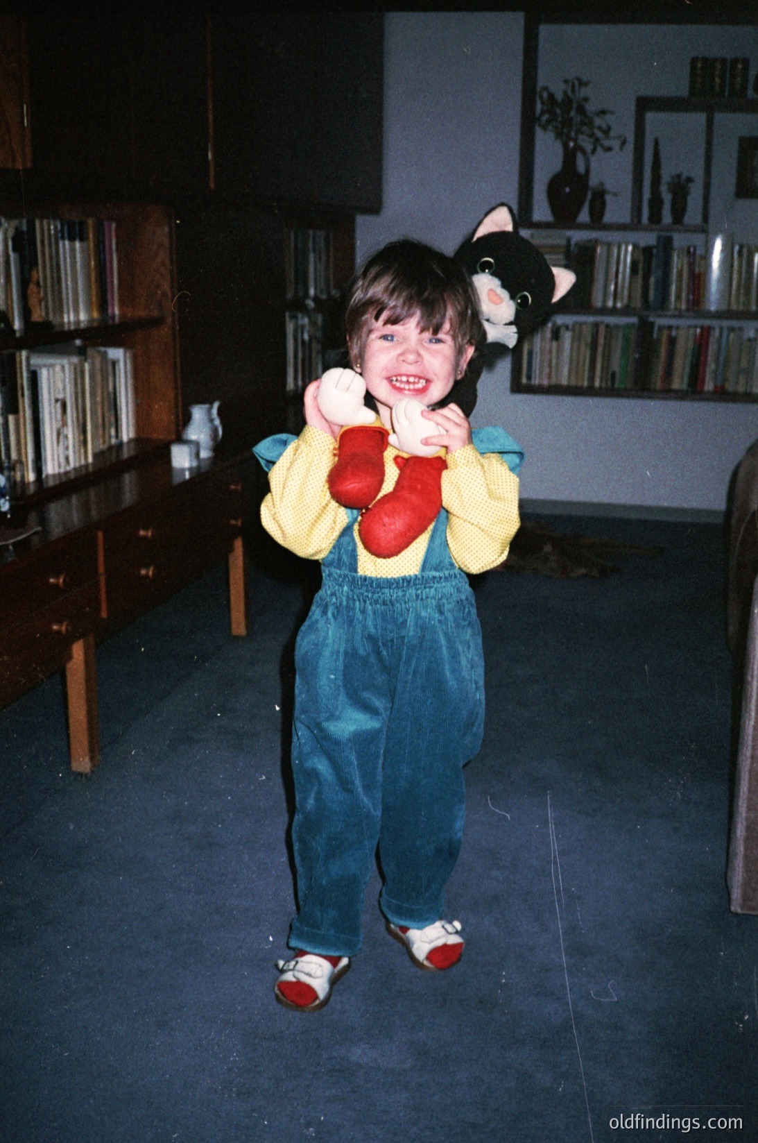Young child in 1990s-style overalls and red mittens poses indoors with a black cat plushie. Wooden bookshelves and vintage furniture suggest a mid-century home library. Warm lighting highlights playful, candid expression.
