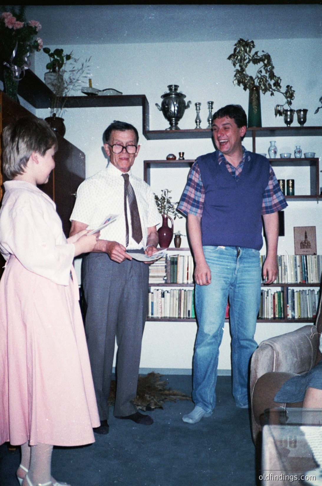 Three individuals in a mid-century living room, likely 1970s–1980s. A man in a striped sweater and jeans stands beside a man in a dress shirt and tie, both smiling. A woman in a knee-length dress holds a book. Shelves display vintage glassware, books, and decorative items. Warm, lived-in interior with floral arrangements.