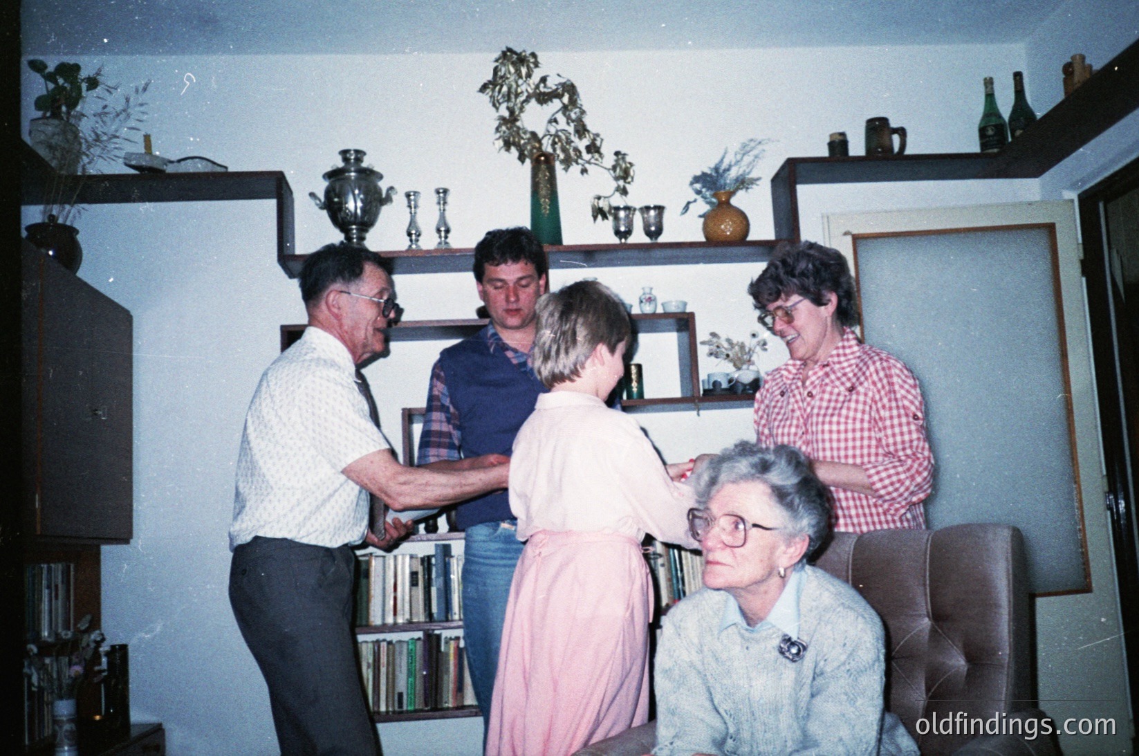 Vintage indoor gathering in a mid-century living room with built-in shelving displaying decorative items like vases, trophies, and books. Five adults pose—men in dress shirts, plaid/checked shirts; women in casual blouses and a floral skirt. Seated woman wears glasses and a lanyard. Retro decor includes a TV on a stand and framed art. Likely 1970s–1980s Western household.