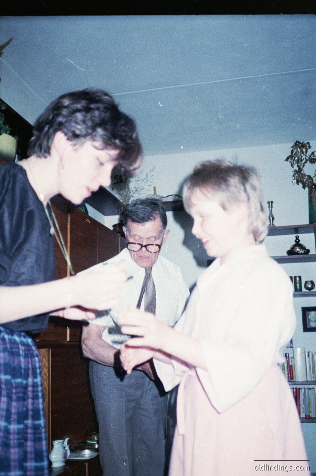 Vintage indoor scene featuring three adults examining a small object, likely a camera or film reel, in a mid-century home. The man in glasses wears a dress shirt and tie, while the woman in a light blouse and skirt leans in closely. The background includes wooden furniture, a bookshelf, and a potted plant. Color tones suggest 1970s–1980s photography.