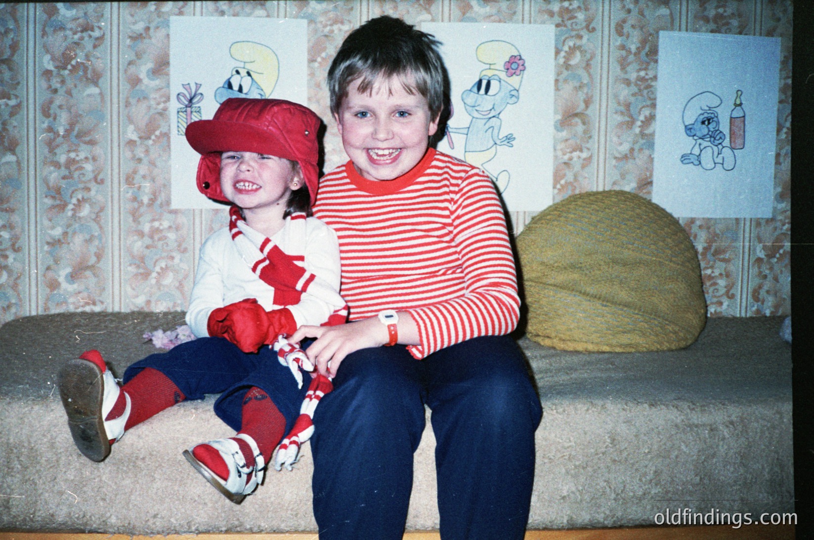 Two children pose indoors on a patterned sofa, likely 1980s–1990s. The boy wears a red-and-white striped sweater, blue jeans, and a watch; the girl sits on his lap in a red hat, striped shirt, and red-and-white sneakers. Smurf-themed posters and a beige pillow frame the scene. Warm, nostalgic family moment.