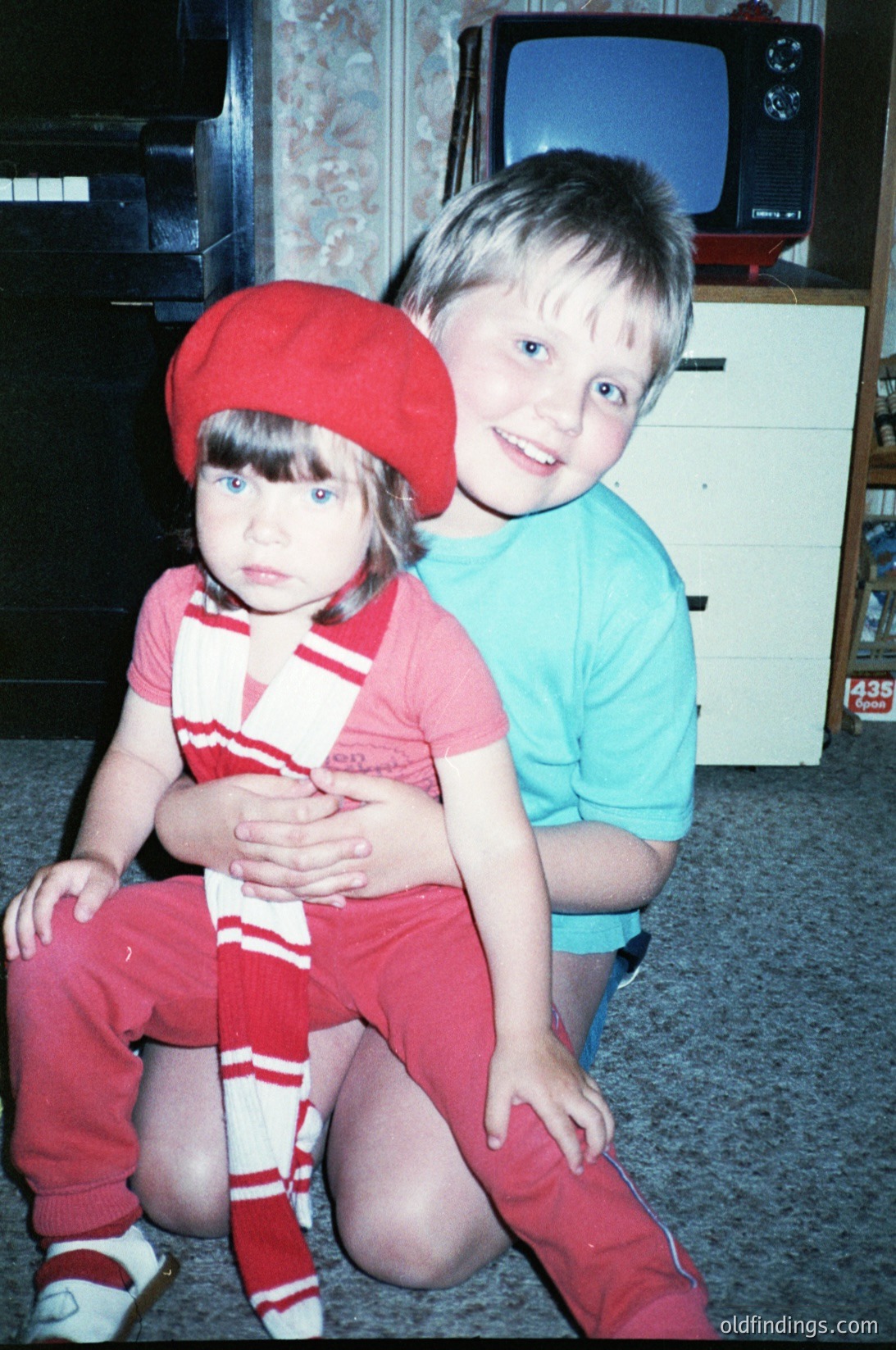Two children pose indoors, likely mid-1980s–1990s. Girl in red beret, striped shirt, and matching pants sits beside boy in teal shirt. Background features a vintage TV, wooden cabinet, and patterned wallpaper. Evokes nostalgic family life.