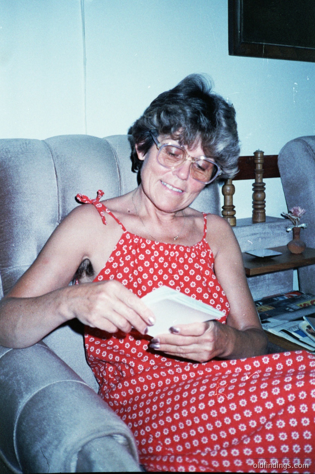 Mid-century indoor portrait of an elderly woman in a floral-patterned sleeveless dress, holding a folded card or letter. She wears round-framed glasses and a red ribbon bow on her shoulder. Background includes a wooden side table with a lamp and scattered papers, suggesting a lived-in 1970s-1980s home setting.