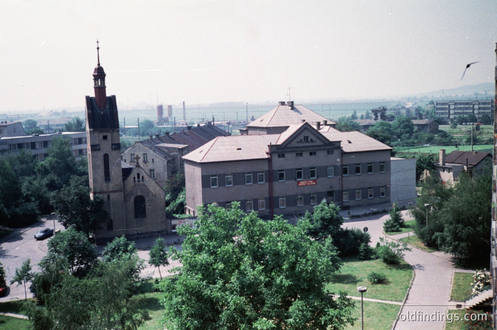 Aerial view of a historic church with a tall brick steeple and arched entrance, adjacent to a mid-20th-century institutional building ( ) --- **Note:** The institutional building’s signage suggests a public or educational function, likely from the 1960s–70s. The landscape and architecture reflect mid-century urban development.