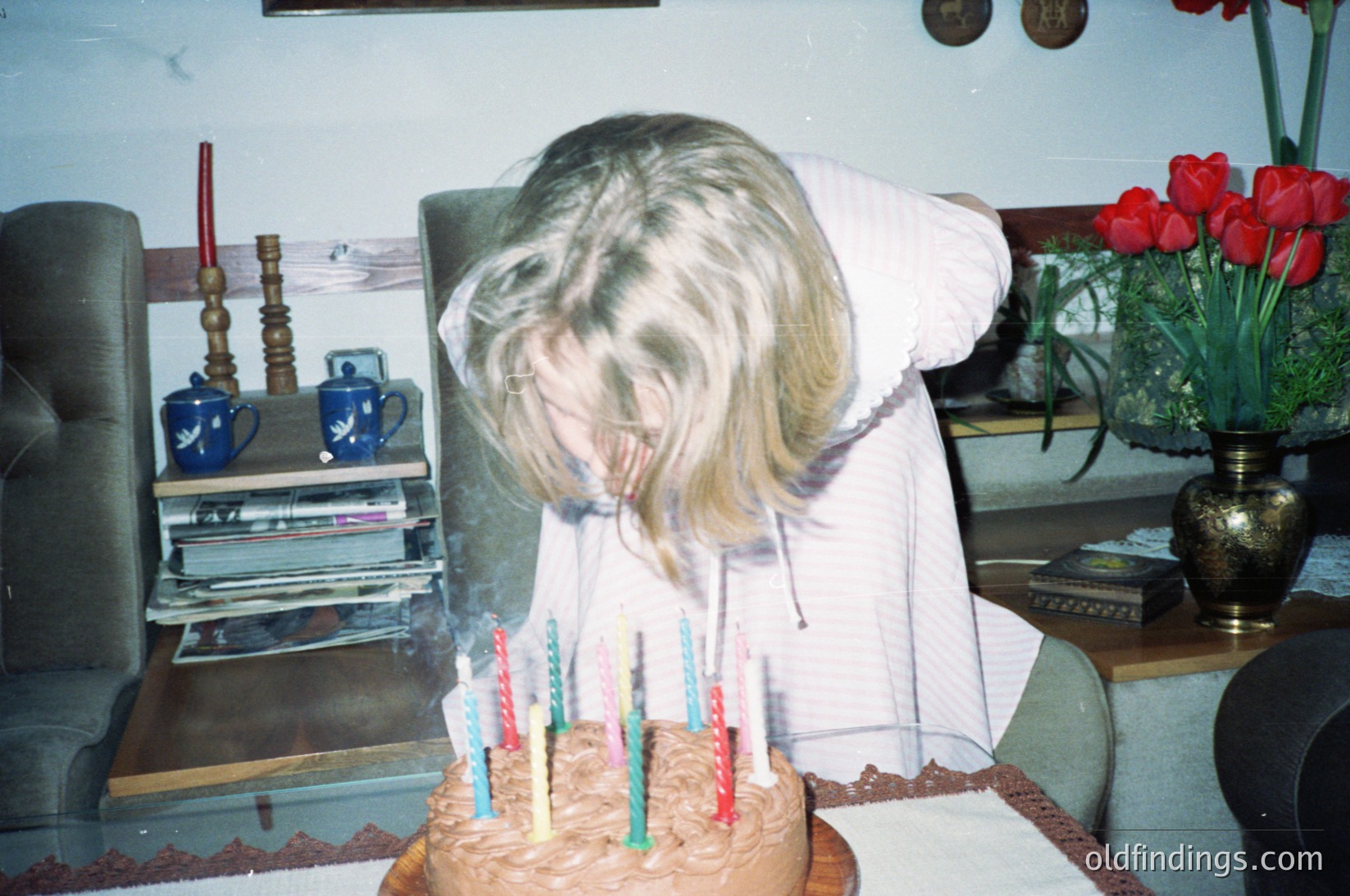 Vintage-style indoor birthday scene featuring a child blowing out candles on a round cake. Soft-focus dog in foreground, white tablecloth, blue mugs, and red tulips in brass vase. Mid-20th century home decor.