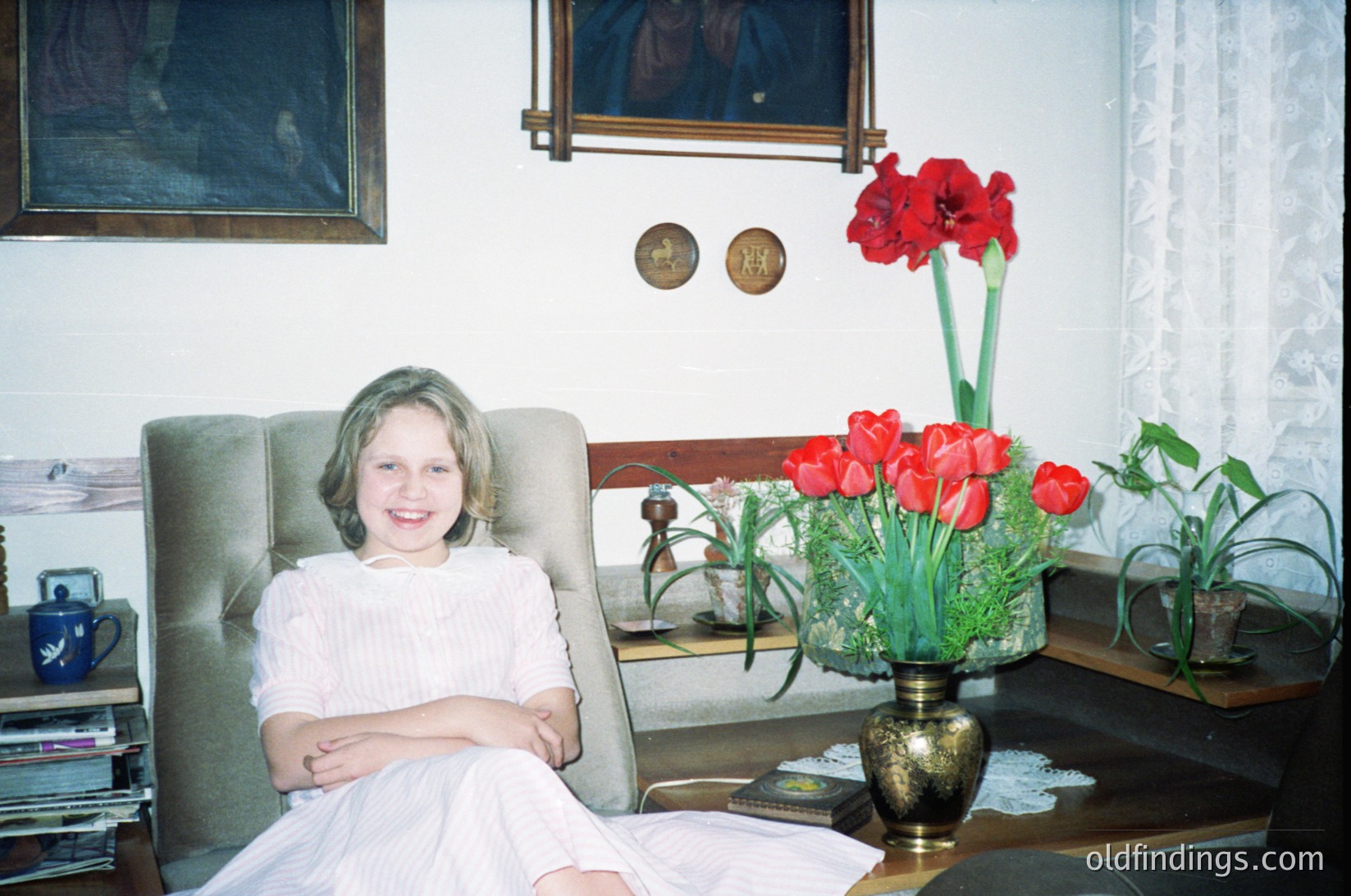 Vintage indoor portrait of a young girl in a white lace dress seated in a mid-century armchair, surrounded by framed art and a vase of red tulips. Decorative medals and a teapot with a floral design add to the 1960s–1970s aesthetic.