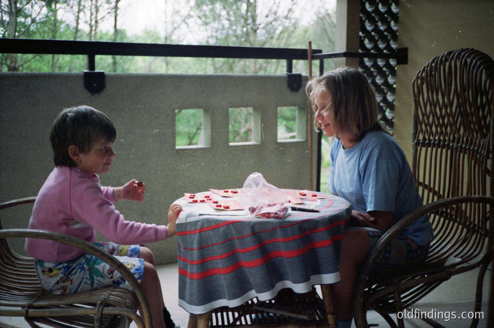Indoor scene featuring two women playing a card game at a small round table draped with a striped blanket. The woman on the left, likely a child, wears a pink long-sleeve shirt and patterned shorts. The adult on the right, seated in a wicker chair, wears a blue T-shirt. The setting appears to be a covered patio or balcony with concrete walls and metal railings. Small framed images are mounted on the wall behind them. [Intimate card game between generations in a cozy patio setting ]