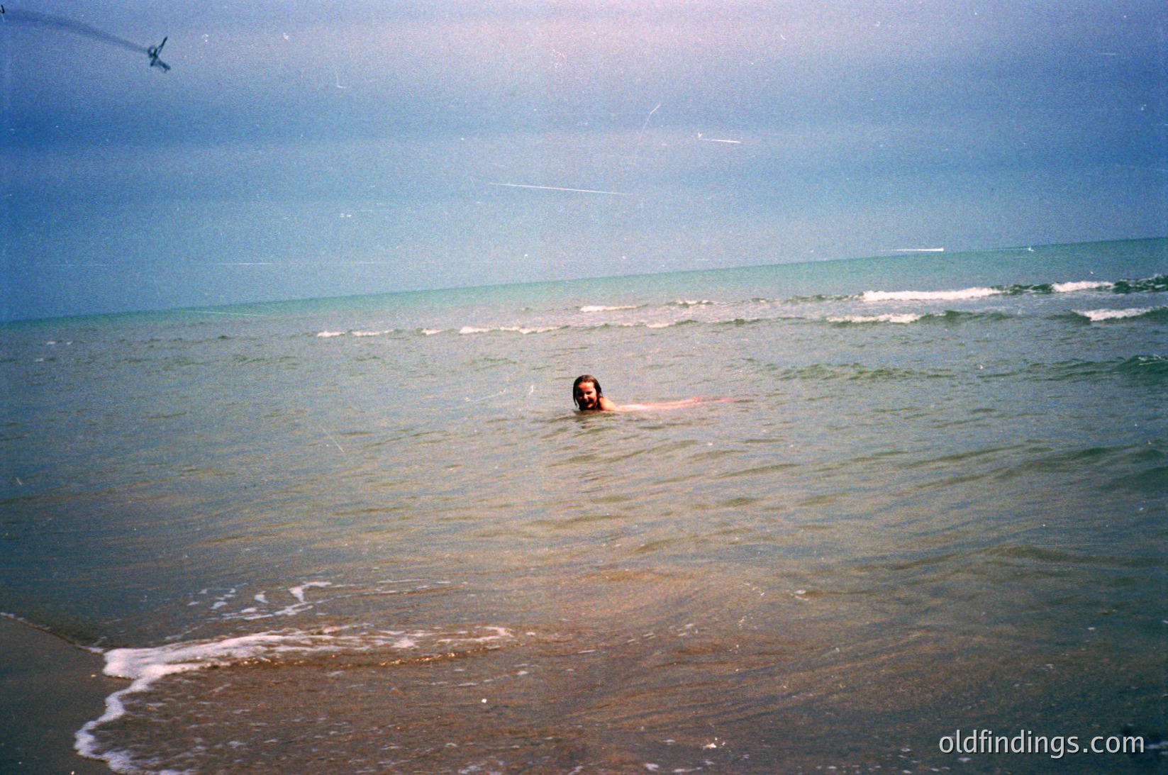 Vintage seaside shot of lone swimmer in calm ocean waves, mid-20th century. Soft focus and muted tones suggest analog film. Gentle waves lap sandy shore under cloudy sky.