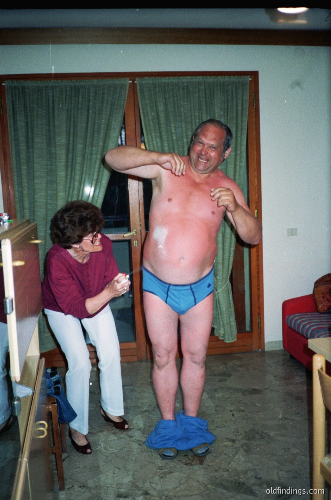 Vintage indoor scene featuring a man in blue undergarments and flip-flops balancing on a single foot, assisted by a woman in a maroon blouse and white pants. Wooden furniture and curtains suggest a mid-20th-century home setting.