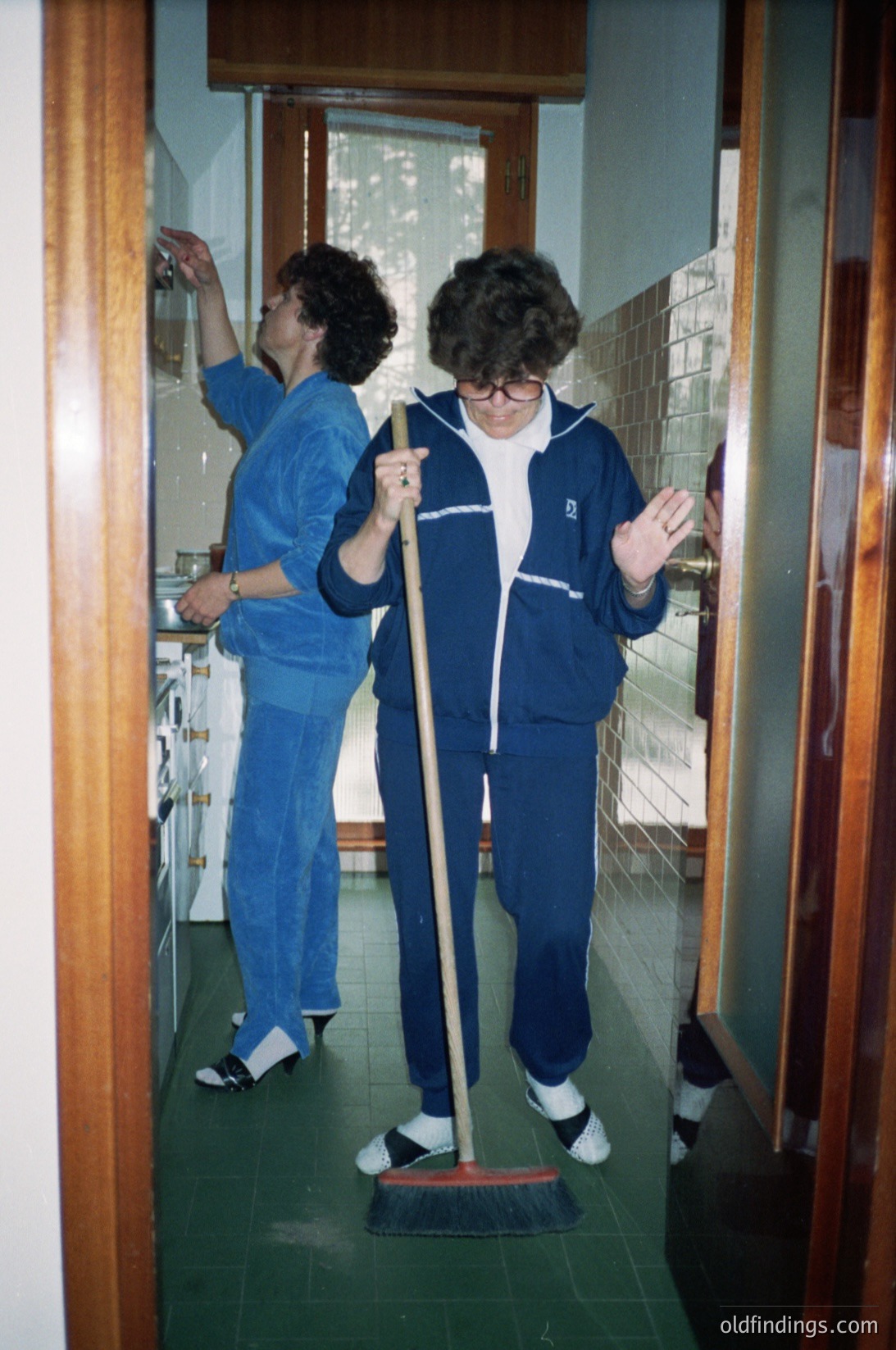 Two individuals in matching blue uniforms mop floors in a retro kitchen, likely from the **1970s-1980s**. The woman on the right wears a track jacket with a visible logo, while the other holds a mop. Reflective glass doors reveal a tiled hallway and white cabinets.