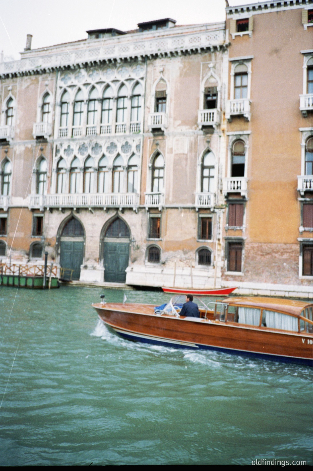 A vintage gondola glides past a weathered Venetian palazzo with Gothic Revival arches and ornate balconies. The waterfront façade shows signs of aging plaster and exposed brickwork.
