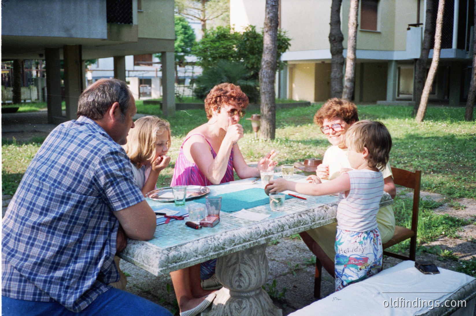 Family picnic scene at a residential courtyard, likely mid-20th century. Adults and children seated at a stone picnic table, engaging in conversation. Adults wear casual 1970s attire (plaid shirt, sleeveless dress), children in patterned shorts and glasses. Lush greenery and palm trees suggest a Mediterranean or Southern European setting. Informal, candid family moment.