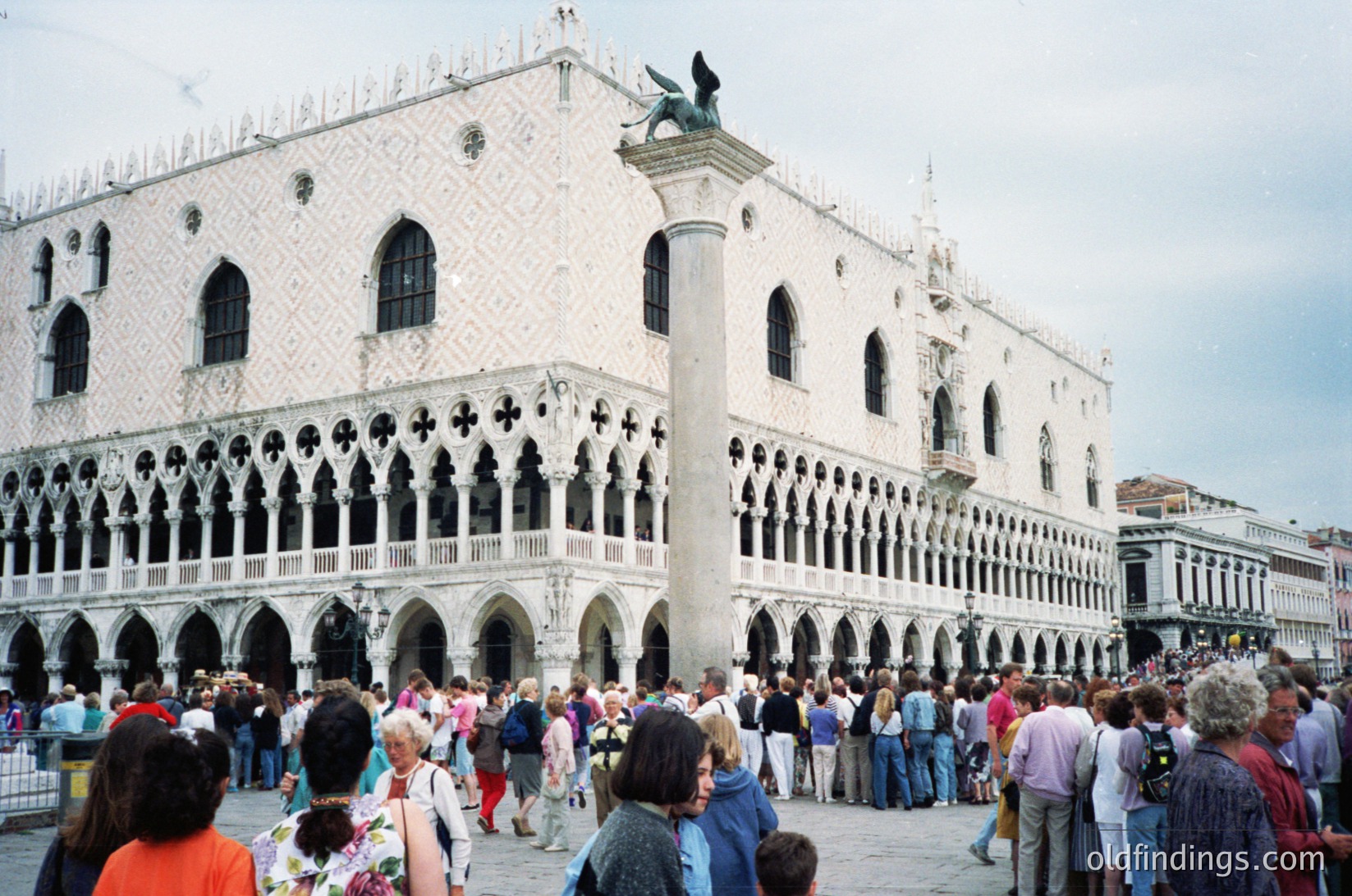 Venetian Gothic architecture of the Doge’s Palace dominates Piazza San Marco, featuring intricate white marble facades with Gothic arches and a prominent winged lion statue atop. Crowds of tourists fill the square, capturing its historic and cultural significance.