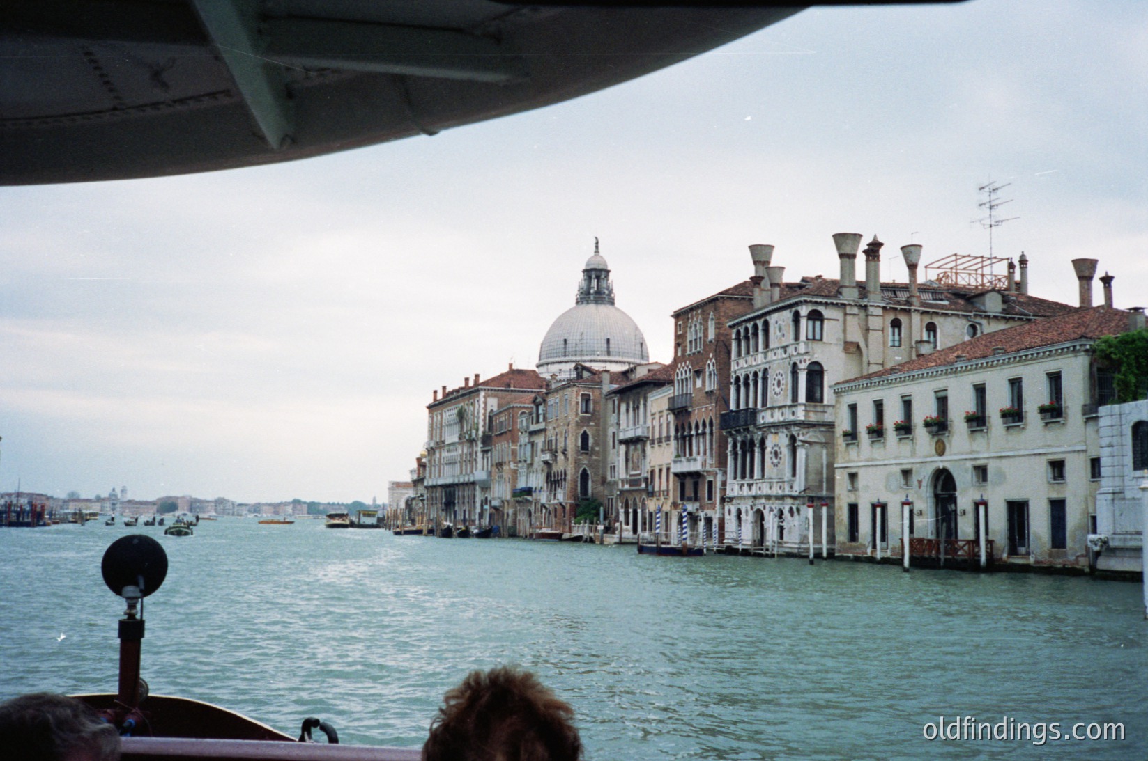 Venetian canal scene showcasing historic palazzos with arched windows and domed church in background. Waterfront architecture reflects 16th–18th century Venetian style. Overcast sky enhances muted tones.