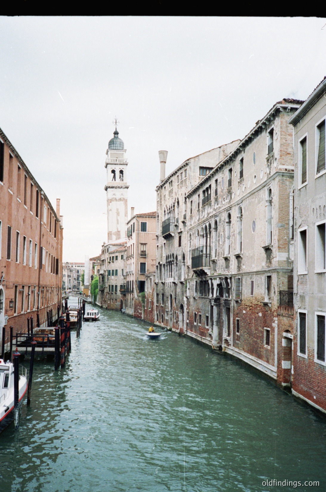 Classic Venetian canal scene with historic architecture. Symmetrical row of 16th–18th century buildings flanking a narrow waterway, featuring arched windows and weathered facades. Distant bell tower (likely Campanile di San Marco) frames the horizon. Overcast sky enhances muted tones. Ideal for travel, architecture, and historical reference.