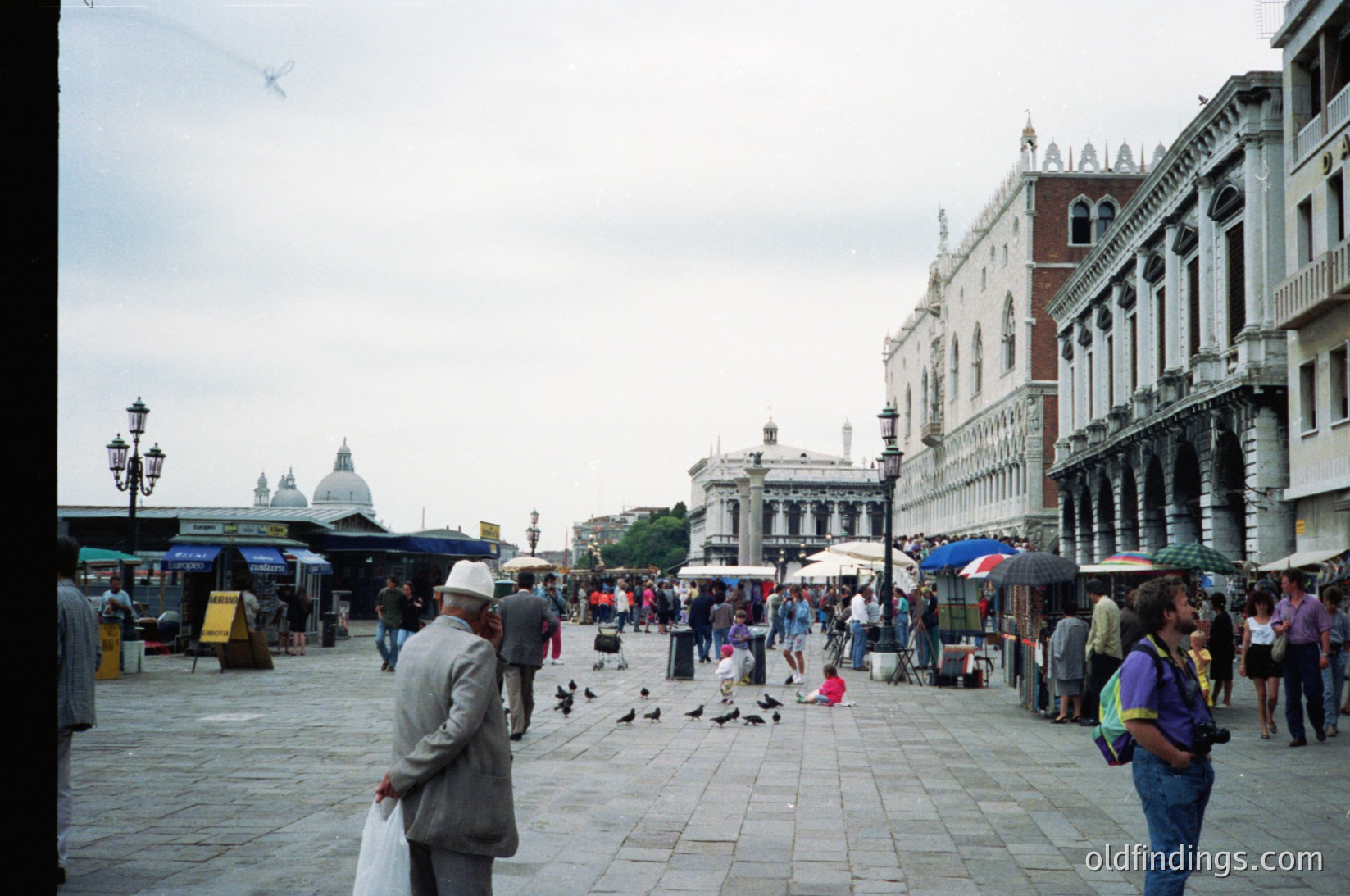 Vibrant Piazza San Marco, Venice, Italy—crowded with tourists and pigeons under overcast skies. Gothic architecture frames the scene, including the Doge’s Palace with its iconic arched windows. Stalls and umbrellas line the square, hinting at a bustling market atmosphere.