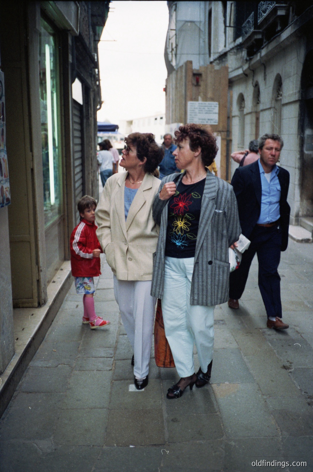 1970s street scene featuring three adults and a child in a European urban setting. Women wear bold patterned blazers and wide-leg trousers; one holds a child in a red dress. Men in suits flank them. Cobblestone alley with shuttered shops and graffiti-style signage.