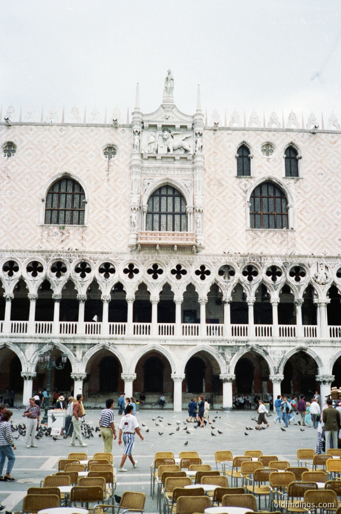 Stunning view of the **Doge’s Palace** (*Palazzo Ducale*) in Venice’s **Piazza San Marco**, featuring Gothic-Renaissance architecture with intricate stonework, arched loggias, and a central clock tower. Yellow folding chairs and pigeons populate the square, reflecting its role as a public gathering space. Likely captured in the **mid-20th century** (1950s–1970s) based on attire and lighting.