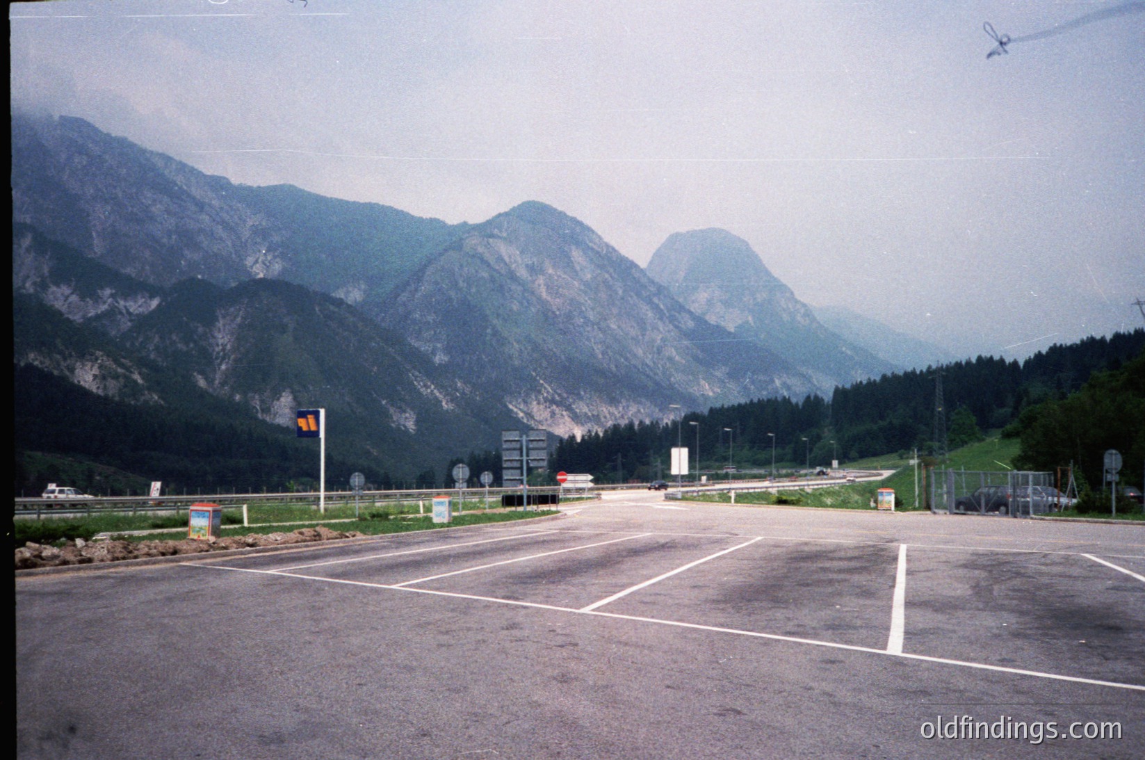 Empty alpine parking lot with forested mountains in background, likely a scenic viewpoint or trailhead. Concrete surface, minimal signage, and distant road. --- *Note: The image lacks distinct landmarks or text for precise location identification, but the alpine setting and style suggest a European mountain region, possibly Austria or nearby. The faded color suggests a 1990s-era photograph.*
