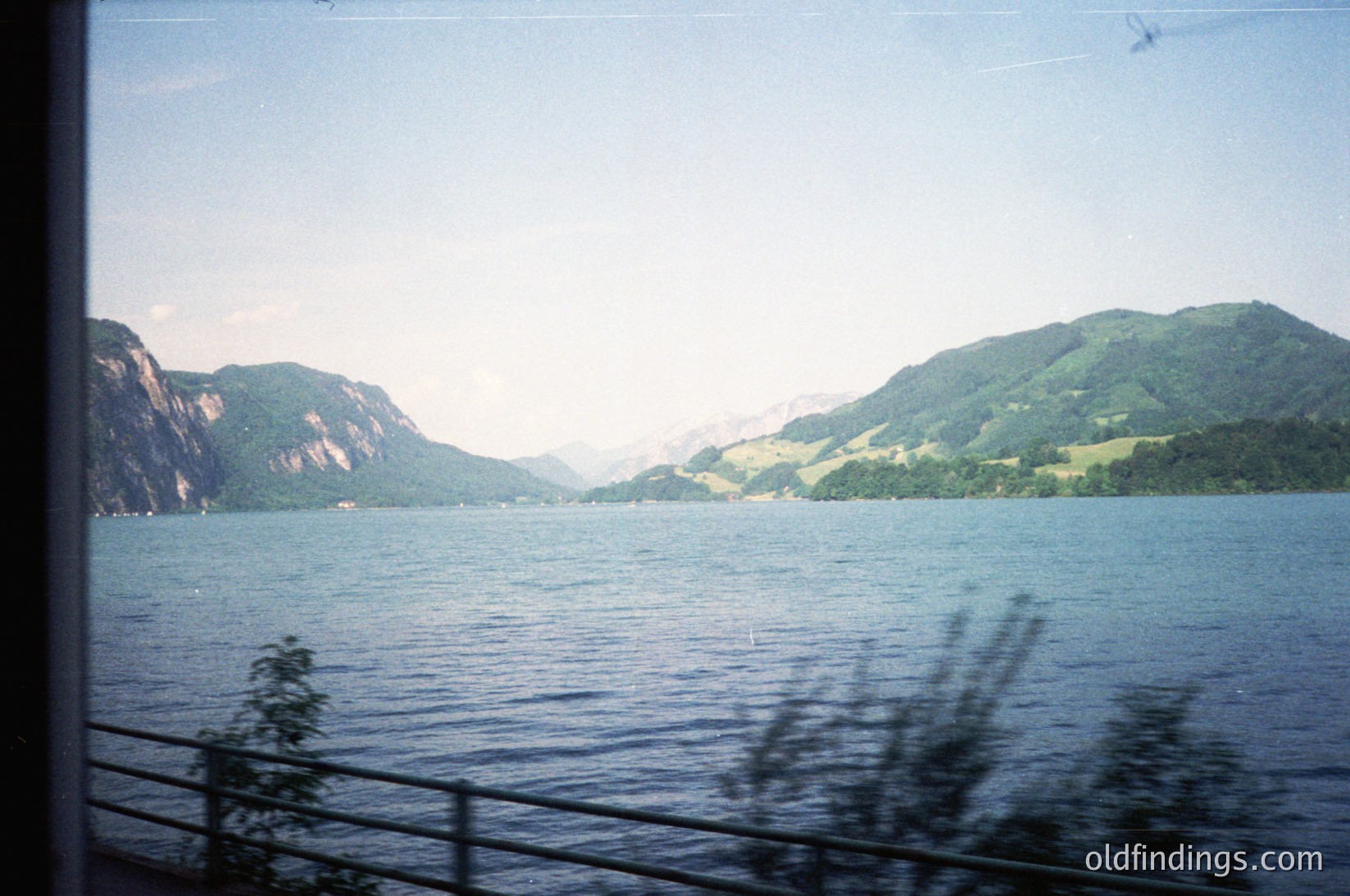 Vintage train window view of a serene alpine lake framed by forested hills, captured through motion blur. Distinctive layered rock formations and misty peaks suggest a Swiss or Austrian lake region.