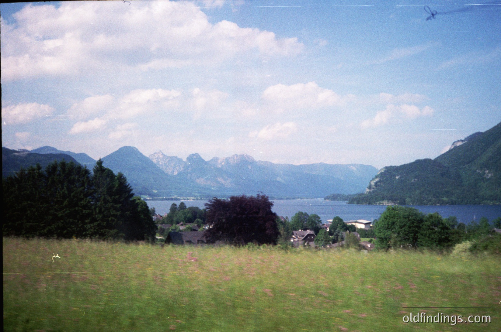Vibrant alpine lake framed by forested hills and jagged peaks under scattered clouds. Mid-ground shows scattered chalets and a narrow road winding along the shore. Soft vintage filter suggests 1980s–1990s European travel photography. Ideal for nature-inspired design or travel inspiration.