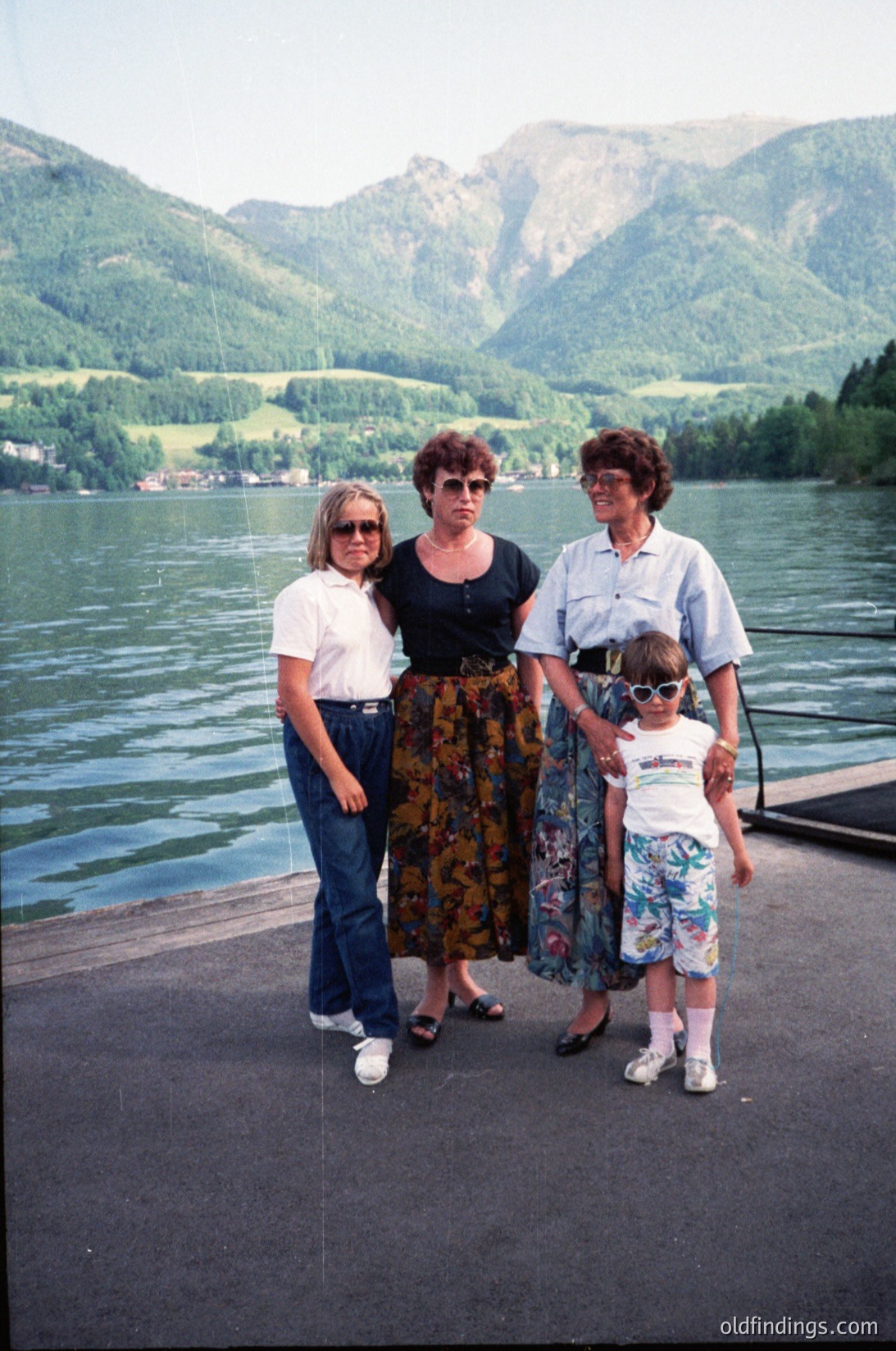 Three women and a child pose on a lakeside pier, framed by alpine scenery. The adult on the left wears a white T-shirt and sunglasses, while the center figure in a floral skirt stands beside a child in a floral dress. The woman on the right, in a light blue blouse, holds the child’s hand. The backdrop features steep forested mountains and a calm lake, suggesting a European alpine lake region, likely the 1980s–1990s.