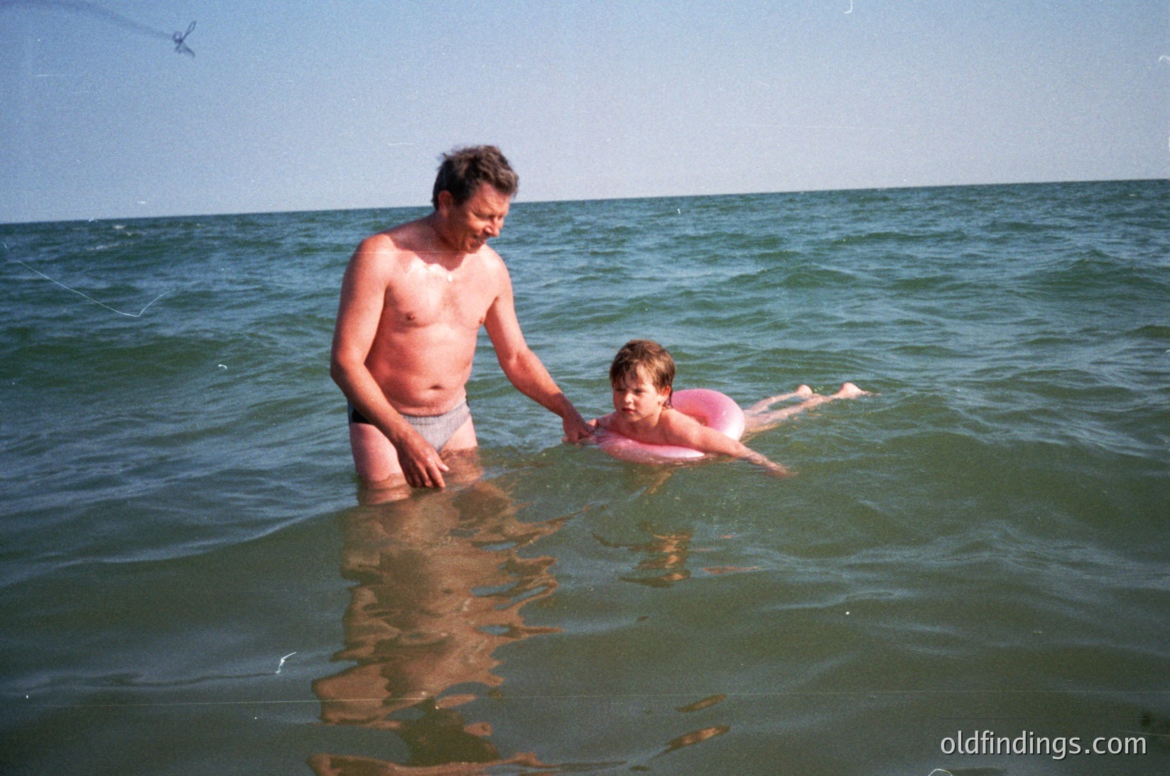 A man assists a young child in shallow seaside waters, mid-20th century. The adult wears loose swim trunks, while the child dons a pink one-piece swimsuit. Waves gently lap against the shore under a clear sky.
