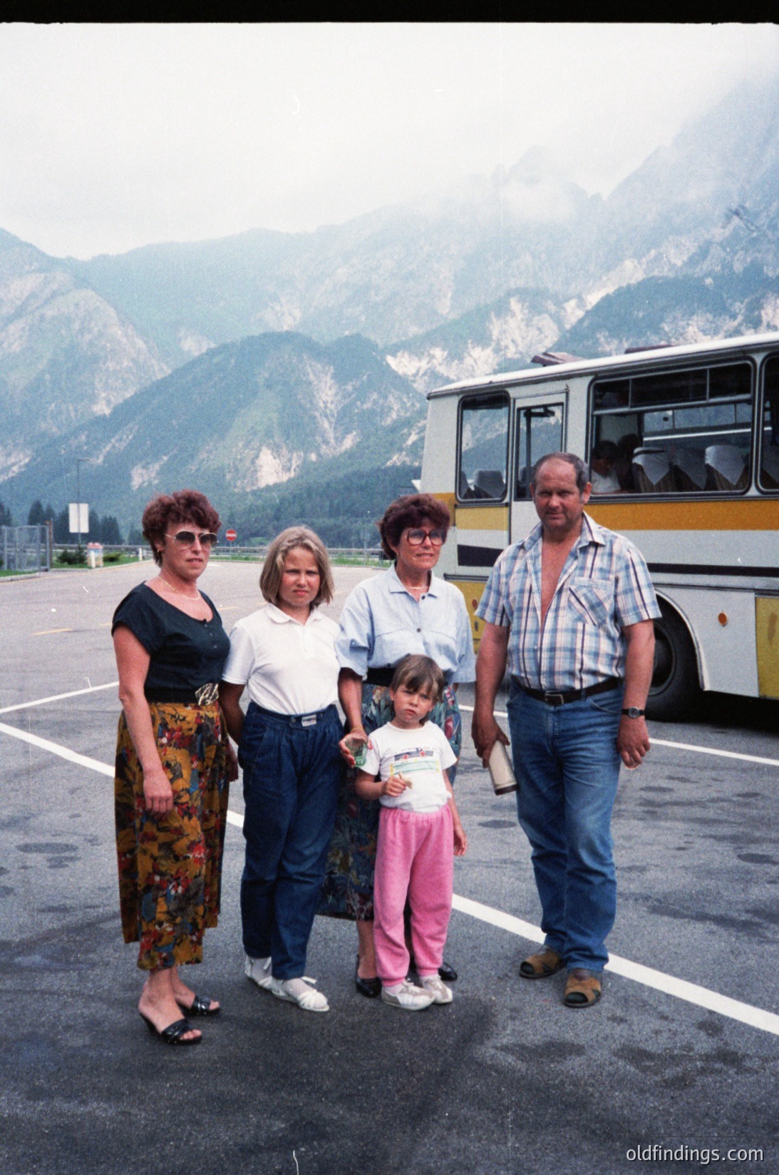 Family group photo in alpine setting, likely 1980s-1990s. Four adults and a young child pose near a yellow-and-blue bus in a paved area surrounded by rugged mountains. Adults wear casual 80s/90s attire: floral skirts, plaid shirts, and jeans. Child wears a pink dress with white polka dots. Bus suggests group travel or tour.