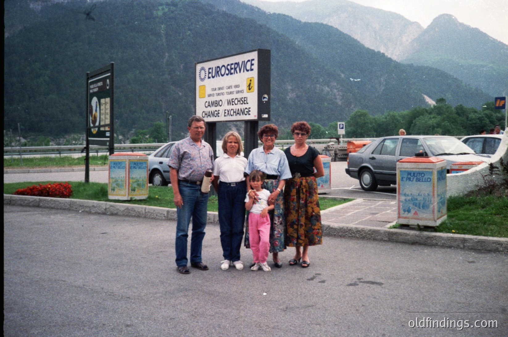 Family portrait at **EuroService currency exchange booth** in alpine setting, likely **1990s**. Four adults and a child pose near signage in German/French ("Cambio-Wechsel/Change-Exchange"). Mountain backdrop suggests European travel hub. Distinctive vintage car and roadside signage enhance nostalgic appeal.