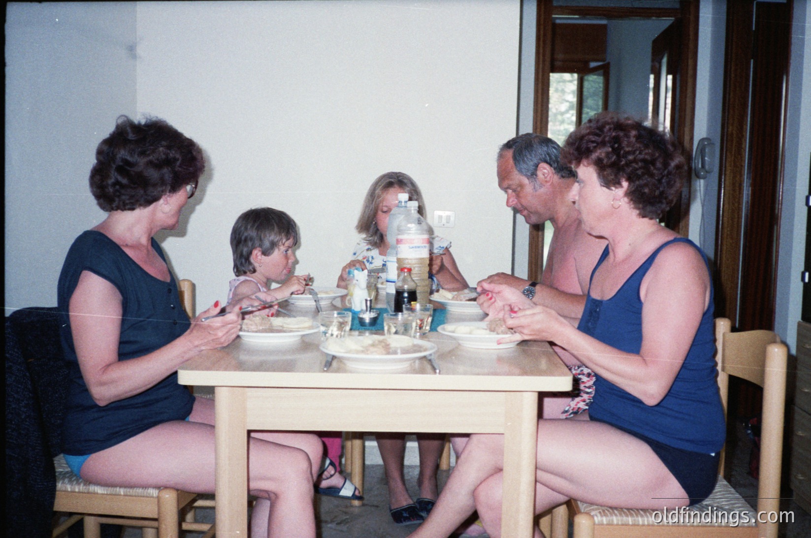 Family meal scene in a casual, sunlit indoor setting, likely a 1970s-1980s European home. Four adults and a child seated around a wooden table, dressed in summer attire (swimsuits, shorts). Bottles, plates, and utensils suggest a relaxed, informal gathering. Lighting indicates natural daylight streaming through windows.