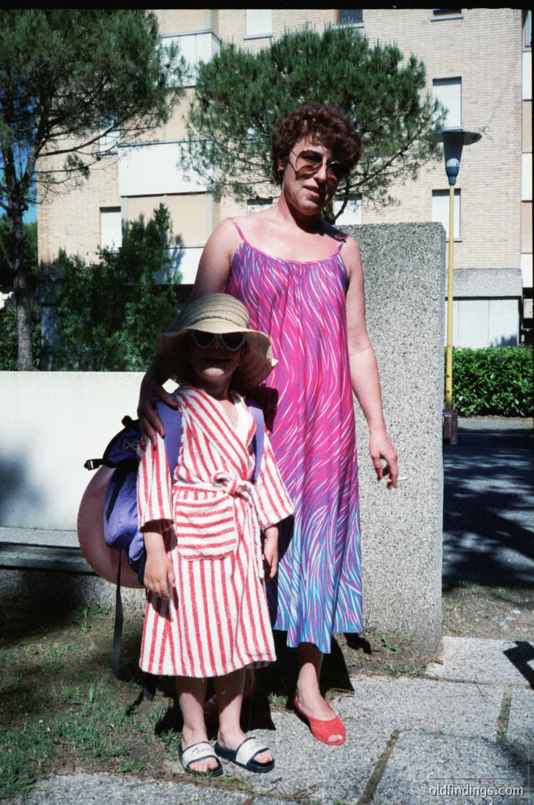 Vintage street portrait: woman in 1970s-style psychedelic dress (pink/blue stripes) and sunglasses poses with child in matching striped dress (red/white) and hat. Urban setting with mid-century concrete buildings and street lamp. Candid, candid lifestyle shot.