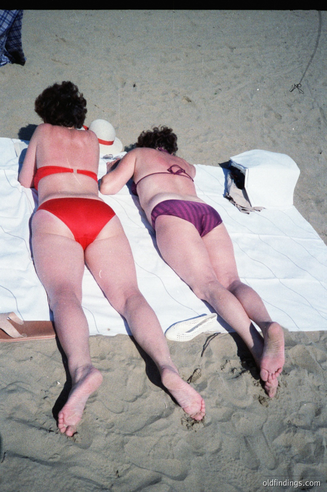 Two women in mid-air on a sandy beach, wearing red and purple bikinis, mid-1970s. Bright sunlight casts shadows on sand; white towels and beach bags visible. Candid, playful moment captures dynamic movement and casual beachwear.