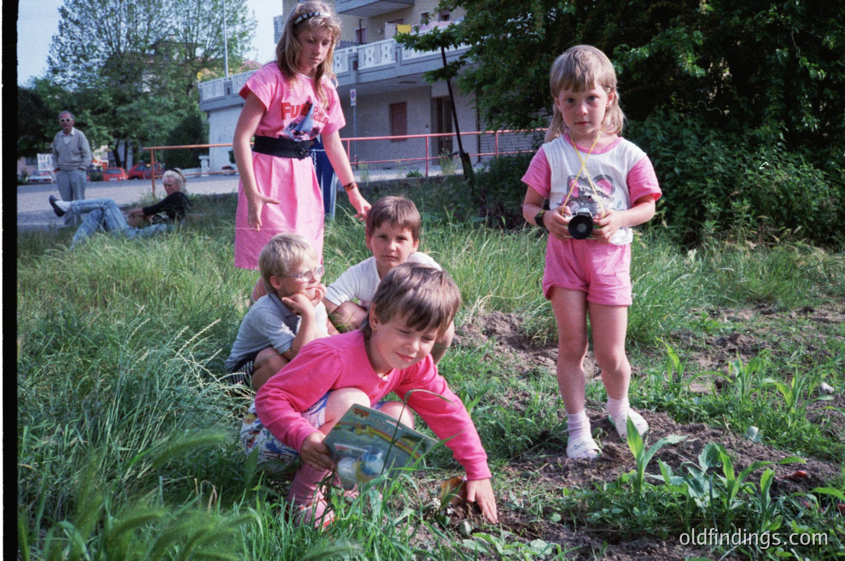 Five children and two adults in matching pink outfits explore a grassy, overgrown area near a residential building, likely a 1970s-80s Eastern Bloc neighborhood. One child kneels with a toy, others pose with cameras. Construction barriers and greenery suggest urban outdoor play.