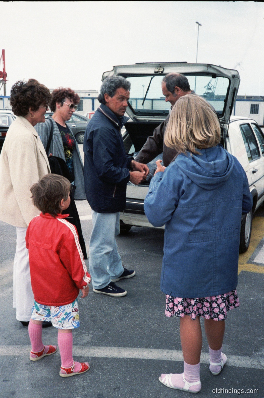 Family group gathers around an open trunk of a vintage station wagon, likely late 20th century. Adults in casual 1980s-90s attire (denim, jackets, sweaters) assist a young child in red jacket and floral shorts. Industrial port or ferry terminal in background with cranes and vehicles.