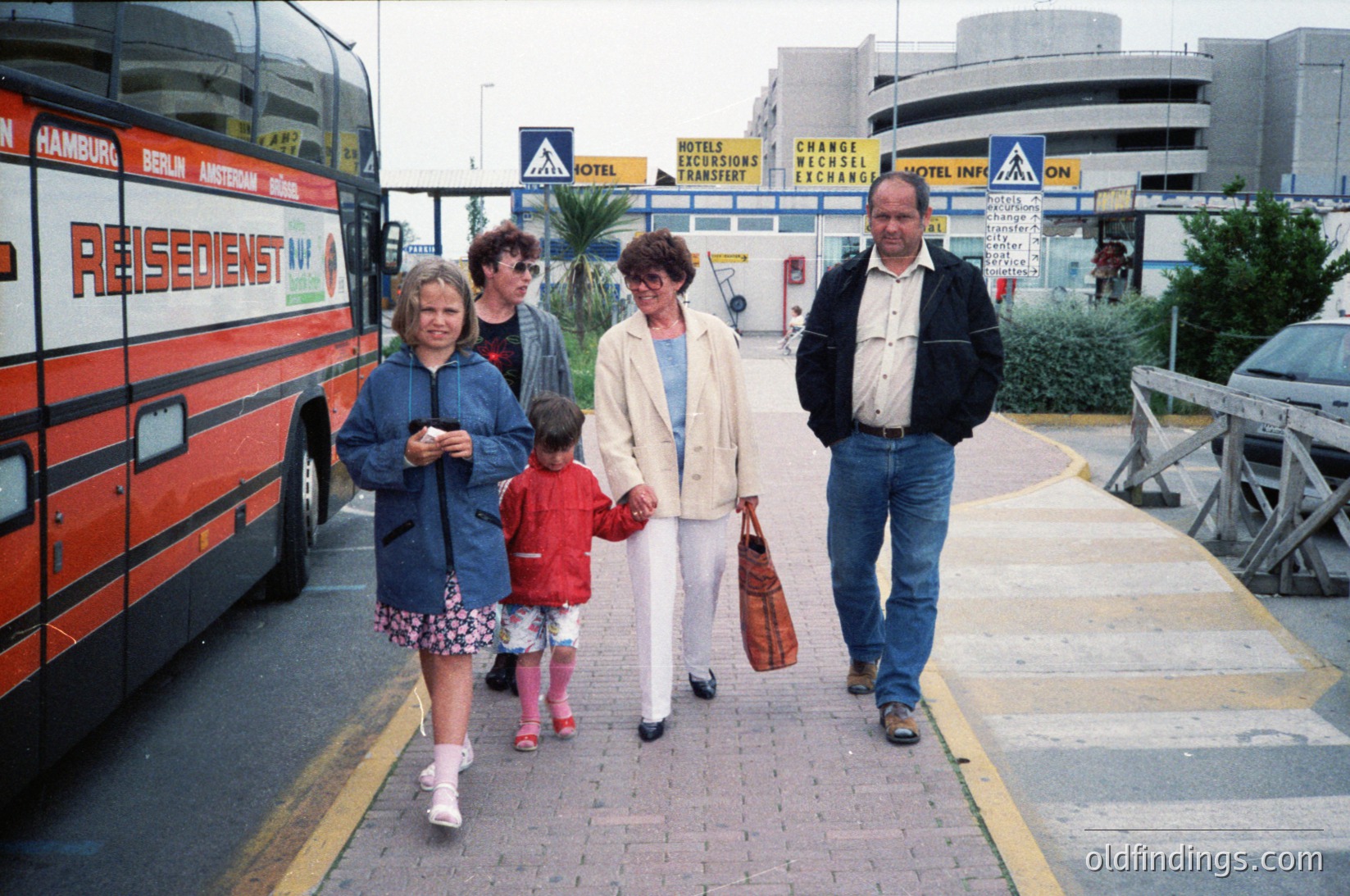 Family group walks near a **Residentur** coach labeled for Hamburg/Berlin, 1990s European travel hub. Signage includes currency exchange, hotel info, and parking directions. Casual attire reflects mid-90s fashion—women in skirts/blouses, men in jackets/jeans. Urban seaside or transit area with palm trees and modernist architecture.