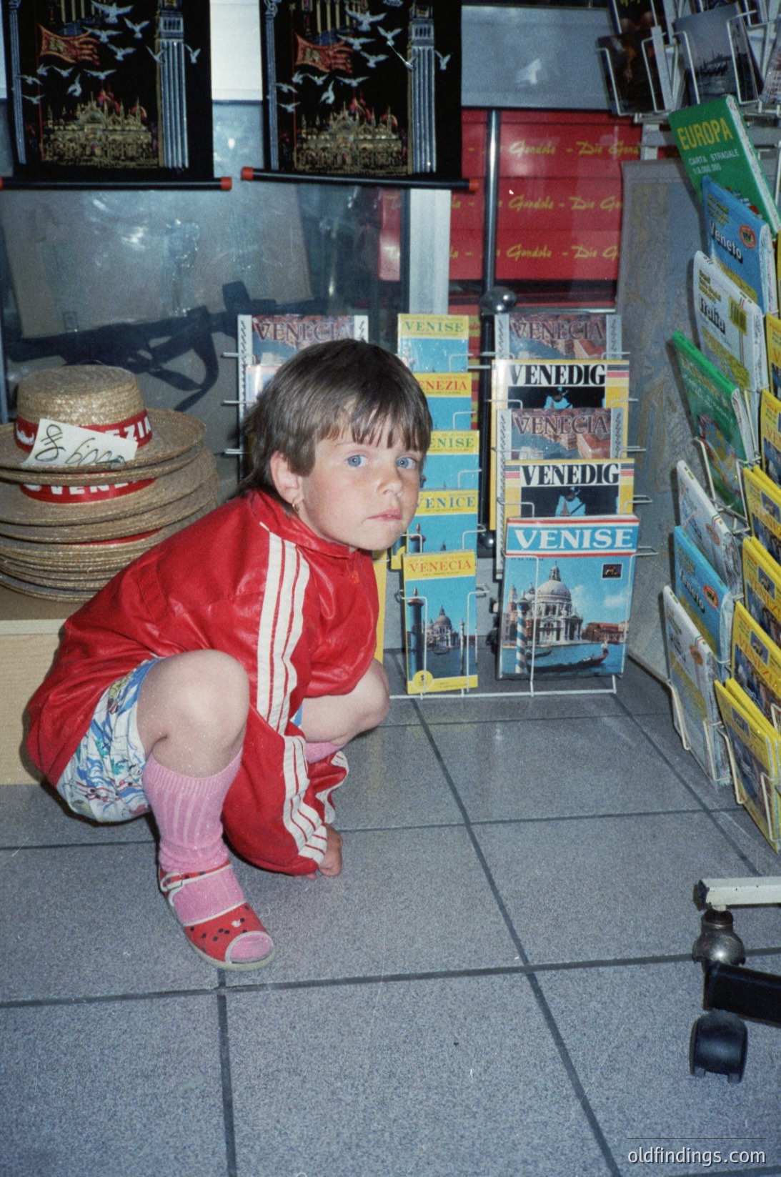 Child in 1980s-style Adidas tracksuit kneeling on tiled floor, surrounded by postcards featuring Venice landmarks. Stacked postcards display "Venezia" and "Venedig" labels. Hat rack with woven straw hats in background.