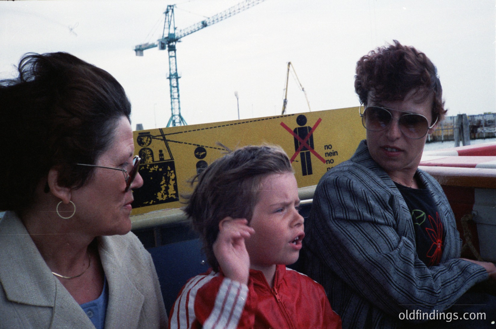 Three individuals stand near industrial construction site with safety signage. Woman on left wears glasses, beige blazer, and gold hoop earrings; child in red striped shirt reacts to sound; woman on right wears sunglasses, striped sweater, and a red patch. Yellow caution banner reads "No entry" with silhouette of worker. Cranes and construction equipment in background suggest mid-20th century urban development.